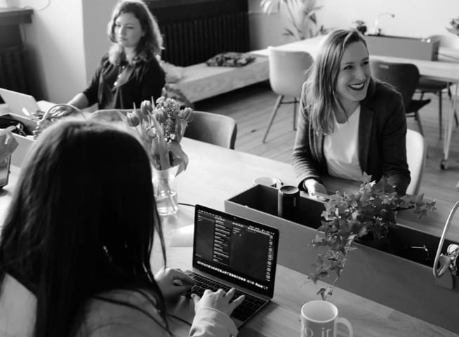 A group of professional looking women all sitting at a table, presumably working, two of them are on laptops.